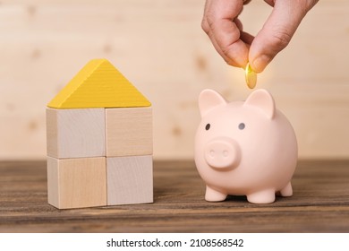 The concept of mortgages and savings for the purchase of housing. A man's hand puts a gold coin in a pink piggy bank standing next to a wooden house made of wooden blocks