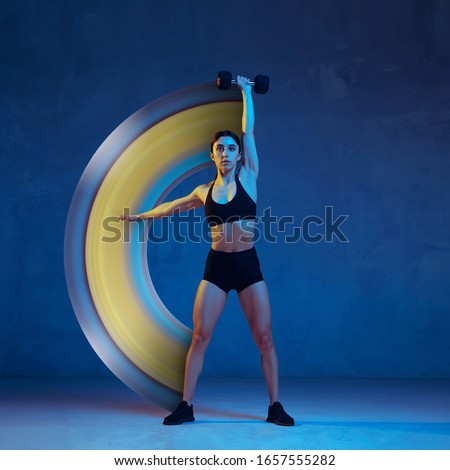 Concept of motion and action in sport. Young female athlete training with the weights on blue background, neon light. Fit and balanced. Sport, healthy lifestyle, movement, wellness. Abstract design.