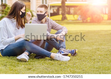 Concept of a new social rules after coronavirus pandemic. Couple students in facial masks sitting on the grass, practicing distance learning, using laptop computer, beside campus building outdoors