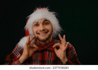 Concept of New Year and Christmas. Minimalistic festive studio background with person. Caucasian guy wears a red hat, a plaid shirt and a bow tie. Emotions of happiness and joy. Front view portrait - Powered by Shutterstock