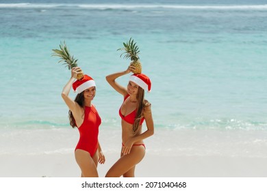 Concept: New Year on the beach. Two beautiful sexy girls  in a red swimsuit and Christmas hats have fund by the sea, hold pineapples in hands over the head