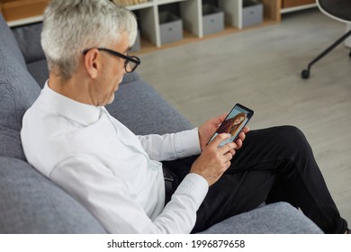 Concept Of Older Adults Using Online Dating Apps Or Websites. Old Bachelor Searching For Future Wife. Senior Man Holding Mobile Phone In Hand And Looking At Photo Of Beautiful Young Woman On Display