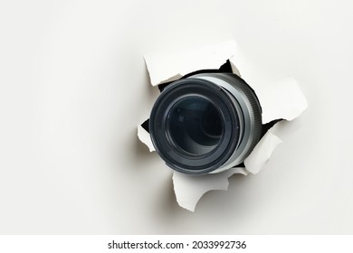 Concept Of Paparazzi Or Hidden Camera, Camera Lens Looks Out Through A Hole In White Paper Wall