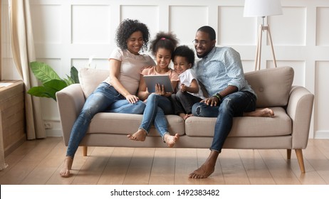 Concept of parental control, modern wireless technology usage, leisure activities at home with children, african full family with daughter and son resting on sofa use tablet computer having fun online