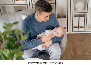 Concept of paternity leave instead of maternity one. Happy young dad holding and feeding newborn baby or infant at home while sitting in armchair in living room. Man holding a little baby on his hands