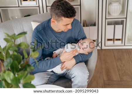 Concept of paternity leave instead of maternity one. Happy young dad holding and feeding newborn baby or infant at home while sitting in armchair in living room. Man holding a little baby on his hands