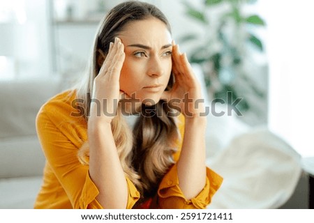 Concept of pensive, tension, and problem-solving. Concerned young caucasian woman in a yellow shirt sitting on a couch at home, pressing her fingers to her temples with a focused worried look away