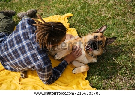 Concept of pet as family member. Young Caucasian man with dreadlocks is resting in park with dog. Male owner strokes German Shepherd on stomach lying on yellow blanket in park. View from above.