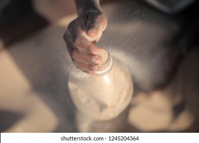 Concept Photography, Eco Building: Aerial Close Up  African Man Hand Filling Up With Sand A Plastic Water Bottle, Making Ecobrick, Outdoors On A Sunny Summer Day, In Gambia, Africa On Atlantic Beach