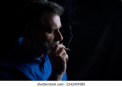 Concept. Portrait Of A Bearded, Stylish Man With Smoke. Secondhand Smoke. Close Up Senior Man Smoking A Cigarette. Handsome Mature Man Smoking Cigarette In Dark Room.