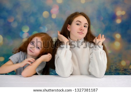 Concept portrait of two cute pretty girls sisters with red hair on a color background smiling and talking. They sit at the table in front of the camera with different emotions and different poses.