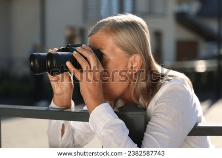 Concept of private life. Curious senior woman with binoculars spying on neighbours over fence outdoors