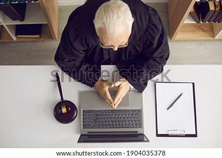 Concept of providing law consultation and legal advice online. Trustworthy wise senior judge, attorney or lawyer sitting at office desk with laptop computer, papers and gavel. High angle, from above