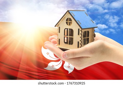 The Concept Of Real Estate Mortgages, Citizenship And Accommodation, As Well As Investment In A Future Home. In Hands Holding A Model Of A Wooden House Against The Background Of The Flag Of Hong Kong.