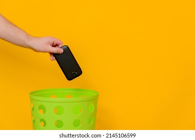 The concept of recycling phones. An old smartphone is thrown into a green trash can for recycling goods, yellow background, space for text