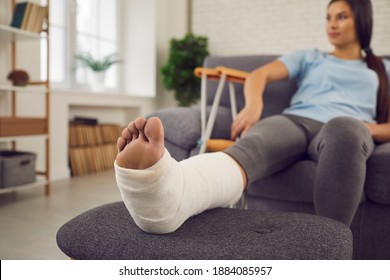 Concept of rehabilitation of people after serious physical accident injury. Female patient with broken leg in plaster cast sitting on sofa. Young woman with foot bone fracture resting on couch at home - Shutterstock ID 1884085957