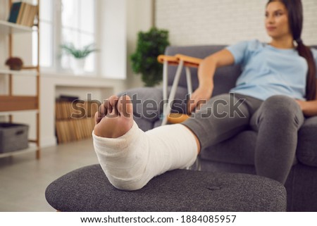 Concept of rehabilitation of people after serious physical accident injury. Female patient with broken leg in plaster cast sitting on sofa. Young woman with foot bone fracture resting on couch at home