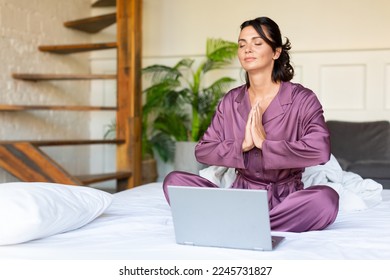 Concept of restoring strength and mental energy through yoga practice - young adult woman meditating in bed in front of laptop monitor. Vertical photo. - Powered by Shutterstock