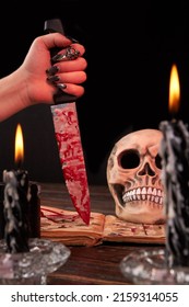 Concept Of Satanic Death Ritual With Blood Sacrifice. Vertical Shot Female Hand Holds Knife Stained With Blood And Wax Over Opened Satanic Bible Book.