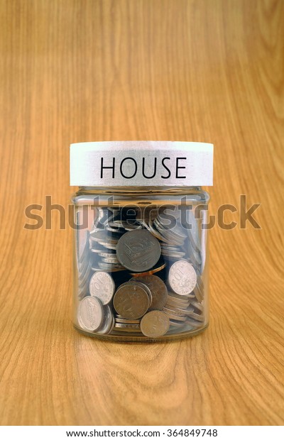 concept of saving, coins in jar with house\
label on wooden\
background.