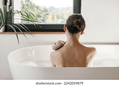 Concept of spa procedure at home. Beautiful woman looking at window while she taking a bath. Girl resting in bathroom, sit in hot water. Back rear view of female washing body with scrubber or sponge - Powered by Shutterstock