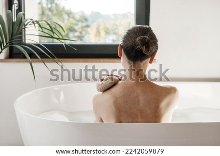 Concept of spa procedure at home. Beautiful woman looking at window while she taking a bath. Girl resting in bathroom, sit in hot water. Back rear view of female washing body with scrubber or sponge