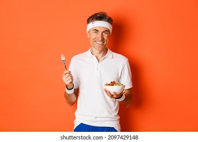 Concept Of Sport, Fitness And Lifestyle. Image Of Handsome, Healthy And Active Male Athlete, Eating Salad And Smiling, Standing Over Orange Background