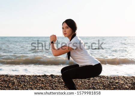 The concept of sport. Young brunette woman doing sports and squats. In the background, the sea and the shore. Close up