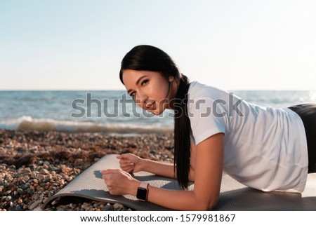The concept of sport. Young brunette woman doing plank exercise on sports Mat. In the background mountains, sea and wild beach. In profile view. Copy space