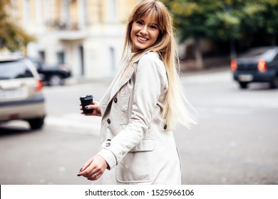 The concept of street fashion. portrait of Young girl dressed in beige trench coat. Posing against the window of the boutique girl smiling and drinking coffee.
