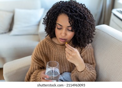 Concept of suffering from terrible pain. Beautiful, sad, upset, unhappy, troubled, weak woman with black hair, wearing casual clothes is sitting on a sofa and taking a pill