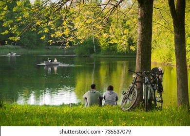 The Concept Of Summer Family Active Recreation. The Couple Sitting Near The Lake With Boats  After A Bicycle Ride, Leaning Their Bicycles To A Large Tree