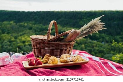 Concept of summer picnic on the hill with amazing view on the green pine forest and hill. Beautiful summer picnic with strawberry, wine and croissants.