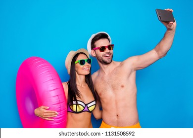 Concept of summer vacation and honeymoon. A guy and a girl dressed in beachwear do selfie on smartphone with an inflatable life ring standing against a blue background - Powered by Shutterstock