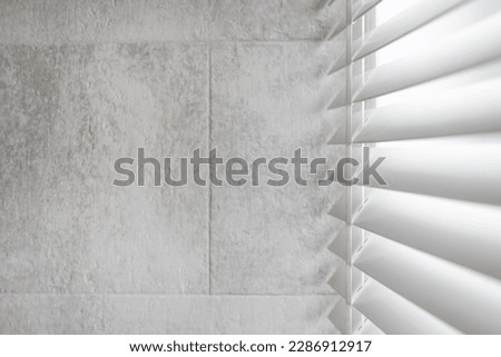 Concept of sunlight and privacy protection on windows in bathroom. Side view of white shutter blinds with wooden slats in tradition venetian style. Copy space on tilled grey wall in loft apartment