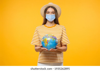 Concept Of Travel After End Of Quarantine. Young Female Tourist Holding Globe With Both Hands, Wearing Medical Mask To Protect From Covid Virus, Isolated On Yellow Background