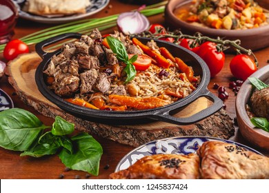 The concept of Uzbek, oriental cuisine. Pilaf in a cast-iron pan, with pieces of lamb lies on a wooden table. Festive table set.