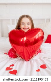 The Concept Of Valentine's Day, A Sweet Child Girl On A White Bed At Home In Red Pajamas Among Hearts Holding A Big Balloon Heart And Smiling With Happiness Congratulating On The Holiday