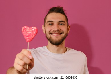 Concept of Valentine's Day. Young Caucasian man with beard shows sweet red lollipop in shape of heart and smiles. Isolated on pink background. Full-face portrait. - Powered by Shutterstock