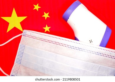 Concept Of Winter Games In 2022 In Beijing, China, Due To Threat Of New Coronavirus Infection. Infrared Thermometer Under Medical Mask On Background Of Red Chinese Flag. Copy Space