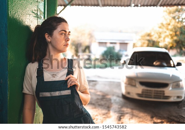 The\
concept of women\'s equality and feminism. A woman in a work uniform\
stands against the wall, holding on to the strap of her overalls.\
In the background, the setting sun and the\
car