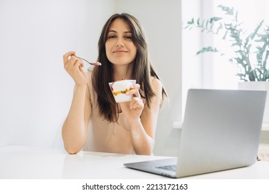 The concept of working at home. Young woman with a laptop in the kitchen, working with pleasure and eating delicious ice cream. - Powered by Shutterstock