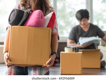 Concept Young Couple Moving House. Asian Woman Holding Box Of Stuff Ready For Relocation And Husband Is Packing Stuff Into Cardboard Box Prepare To Move.