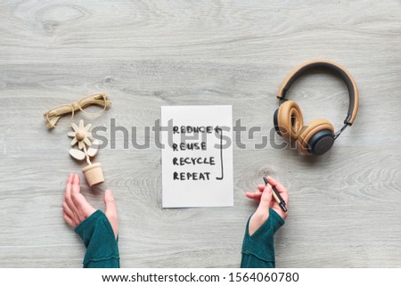 Concept zero waste concious consumption flat lay, Top view of hands holding trendy wooden flower, moleskin notebook with cover from craft cardboard, reading glasses and earphones on wooden table