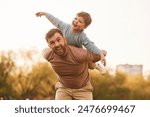 Conception of imagination, sitting on shoulders and flying. Happy father with son are having fun on the field at summertime.