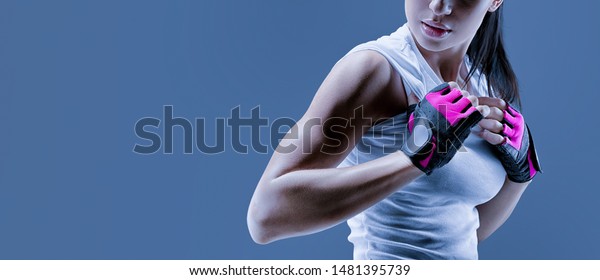 Conceptual close up portrait of fitness athletic young\
female model in sports clothing showing her well trained body\
Confident female bodybuilder with power hand in gloves over toned\
blue background 
