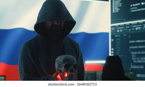 Conceptual image of a hooded russian hacker creating hazardous alerts, holds a skull and warning of death threats. Russian federation works on cybercrime and violence, panic and torment. Camera A.