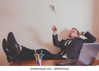Conceptual image: professional burnout, laziness, unwillingness to work. Slacker businessman sitting in office with legs on the table and playing with paper plane. 