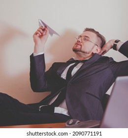 Conceptual image: professional burnout, laziness, unwillingness to work. Close up portrait of slacker businessman sitting in office with legs on the table and playing with paper plane. 