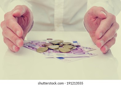 Conceptual Image Of Protecting The Euro Or Your Savings With A Businessman Cupping His Hands On Either Side Of A Pile Of 500 Euro Banknotes And Coins Safeguarding His Investment And Nest Egg.